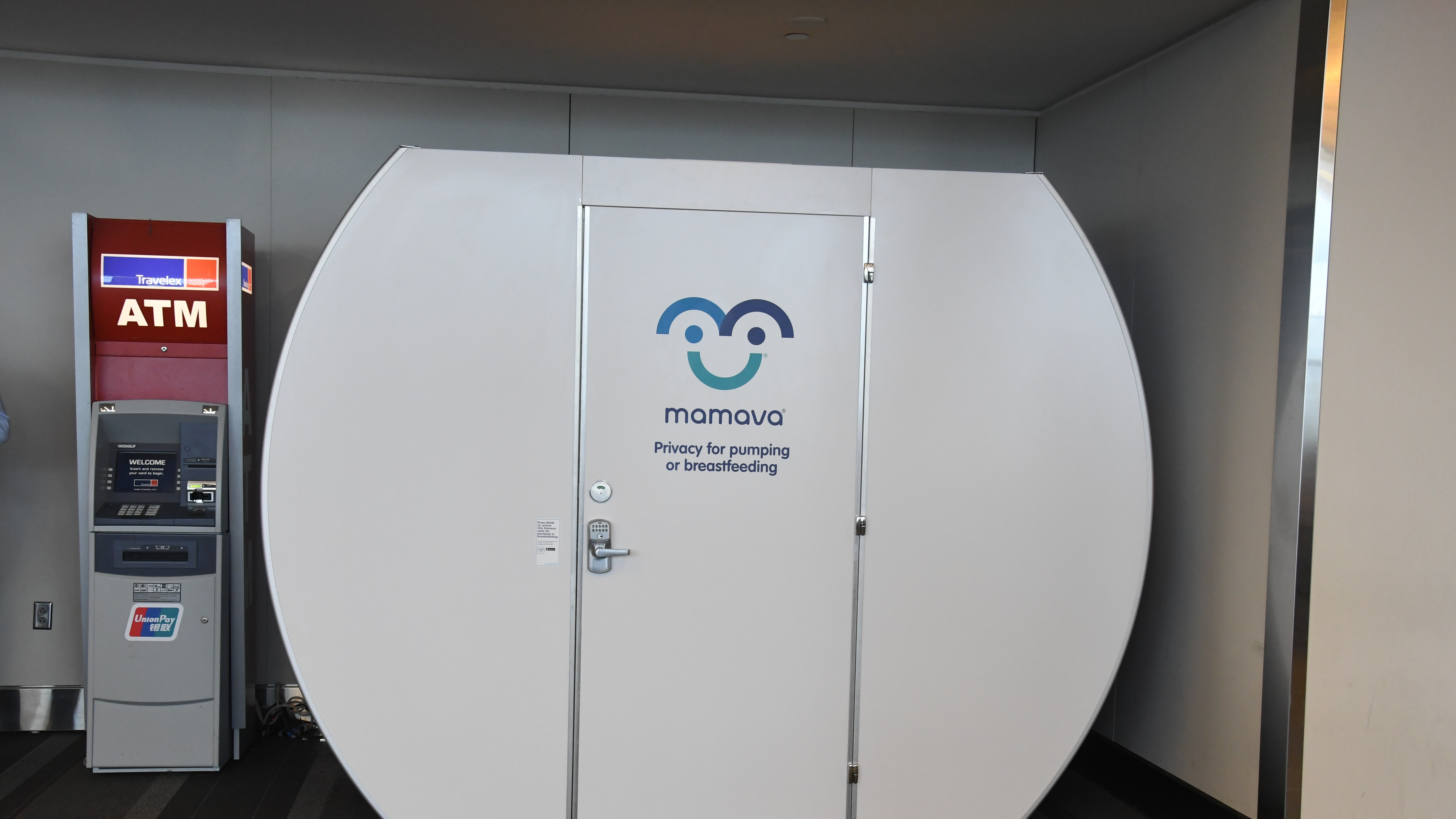 A Mamava lactation pod is situated in an indoor space next to an ATM. The pod is white with a logo and the words "Privacy for pumping or breastfeeding" on its door.
