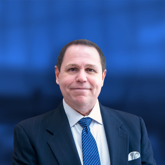 Professional headshot of a man in a dark suit with blue tie and pocket square against a blue background