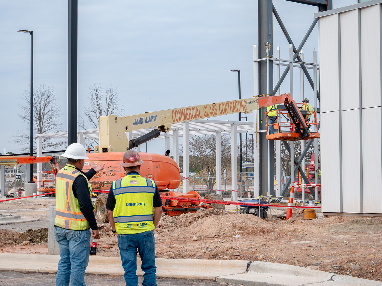 Construction workers in safety vests observe a JLG lift and orange boom lift at a commercial building site.