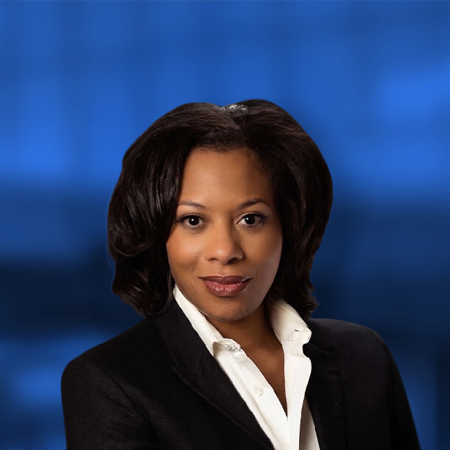 Professional headshot of a woman with dark hair wearing a black blazer and white shirt against a blue background.