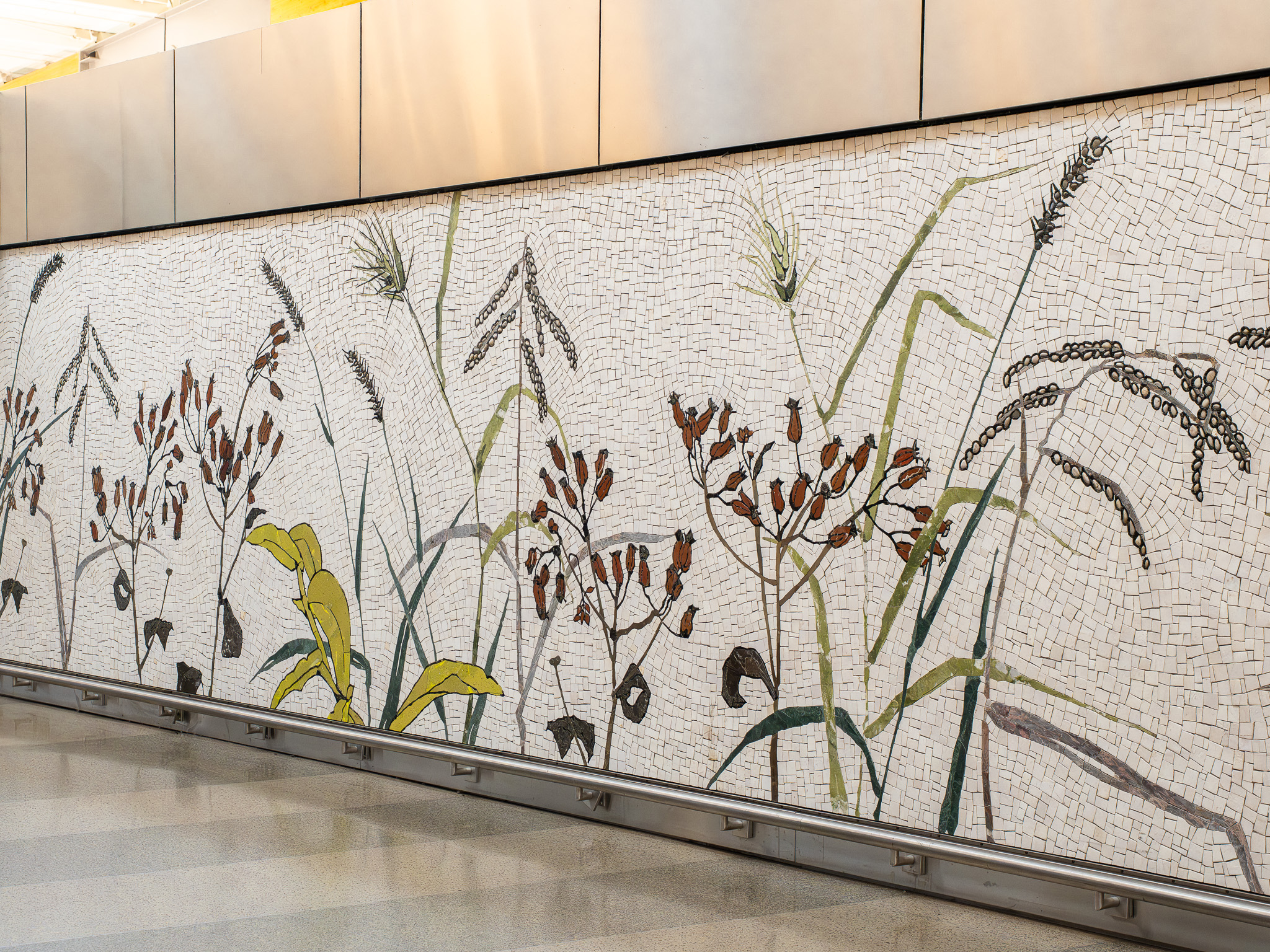 Mosaic wall mural depicting native grasses and plants in brown, green, and yellow tones along a wall.