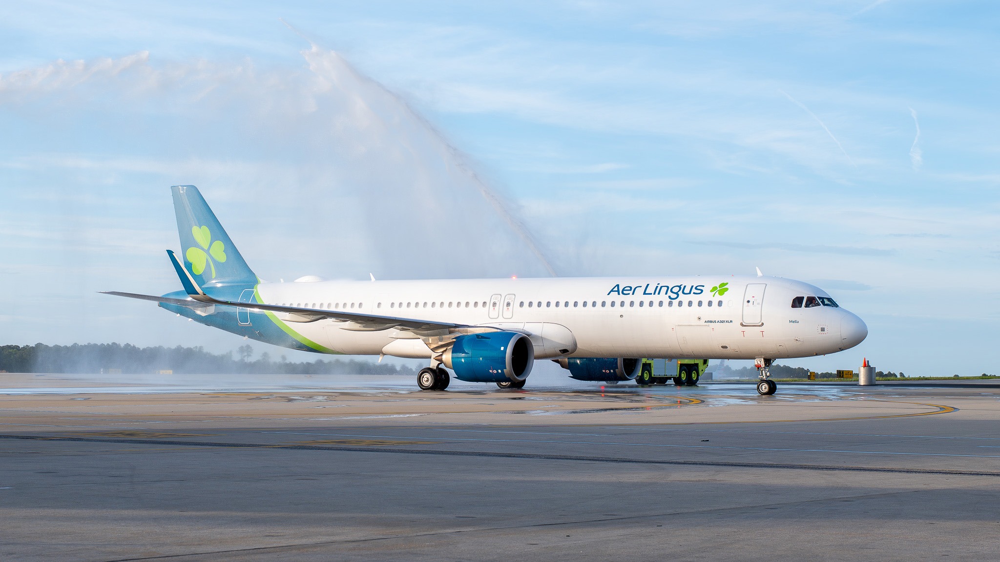 Aer Lingus airplane on a runway being greeted with water cannons, against a clear sky backdrop.
