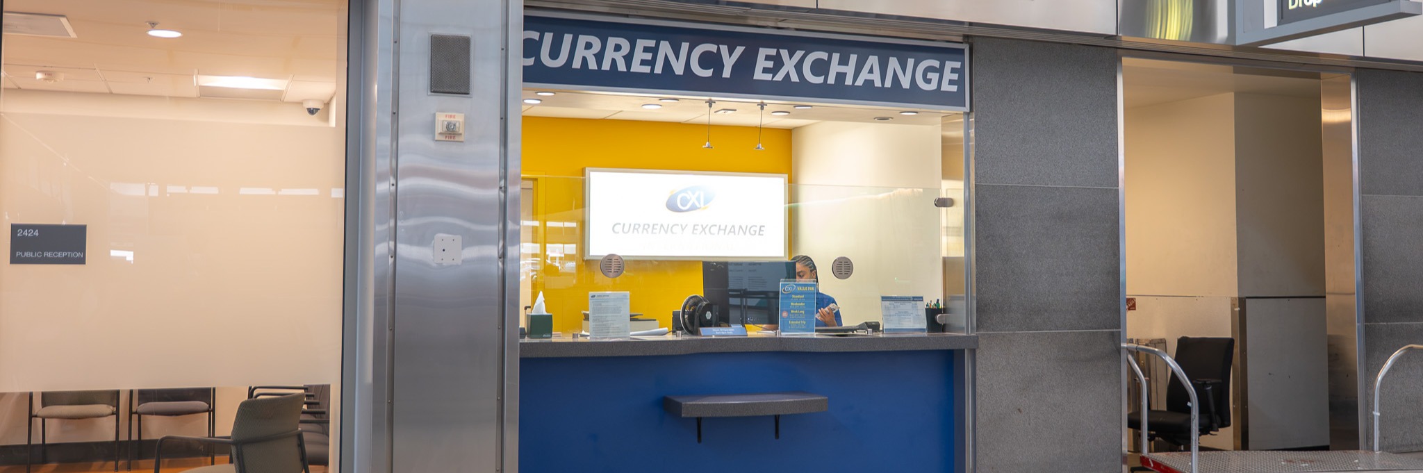 CXI Currency Exchange International kiosk in an airport with blue counter, yellow interior walls, and service window.