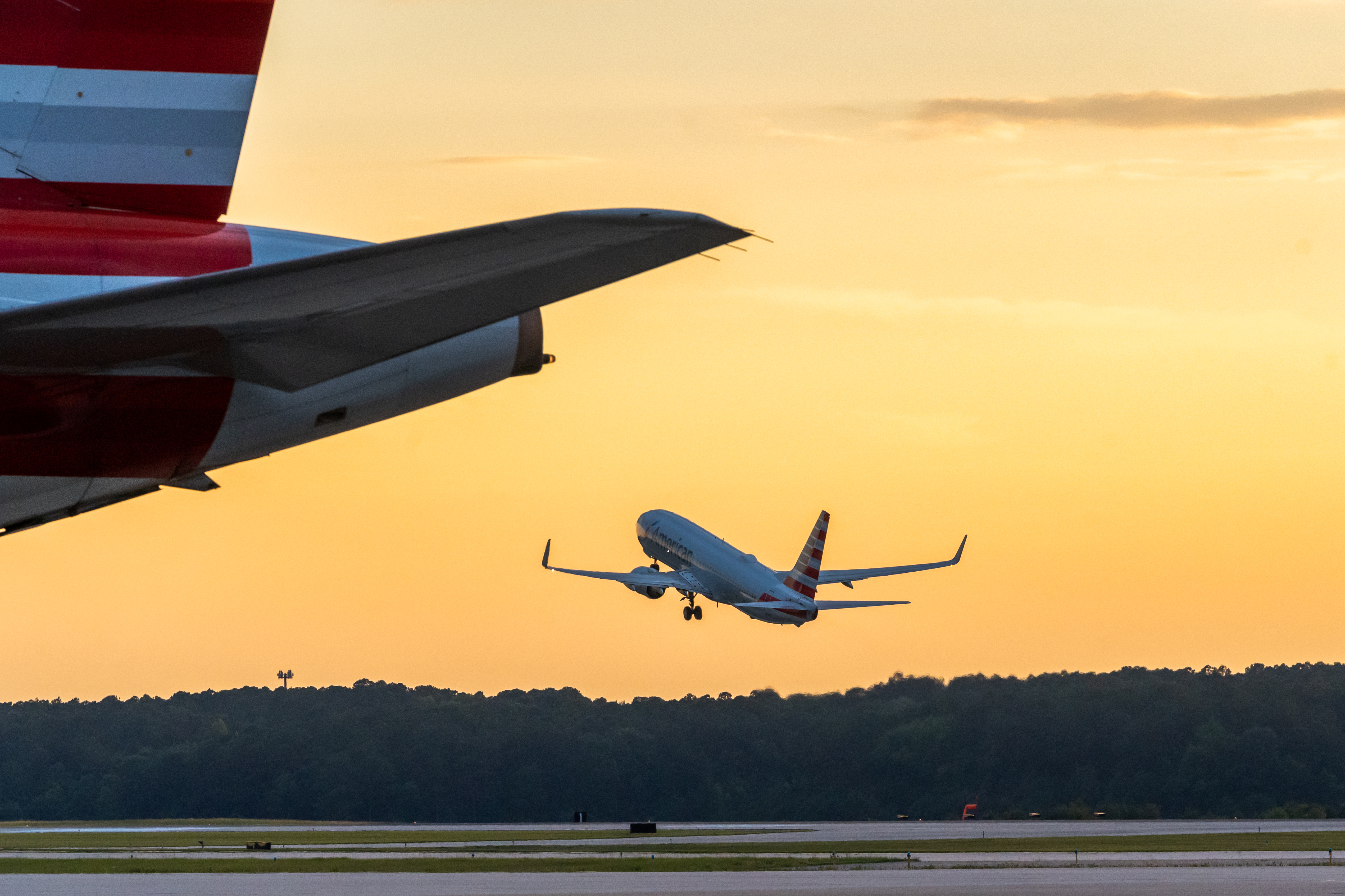 Commercial airplane taking off at sunset with another aircraft's wing visible in the foreground against a golden sky.