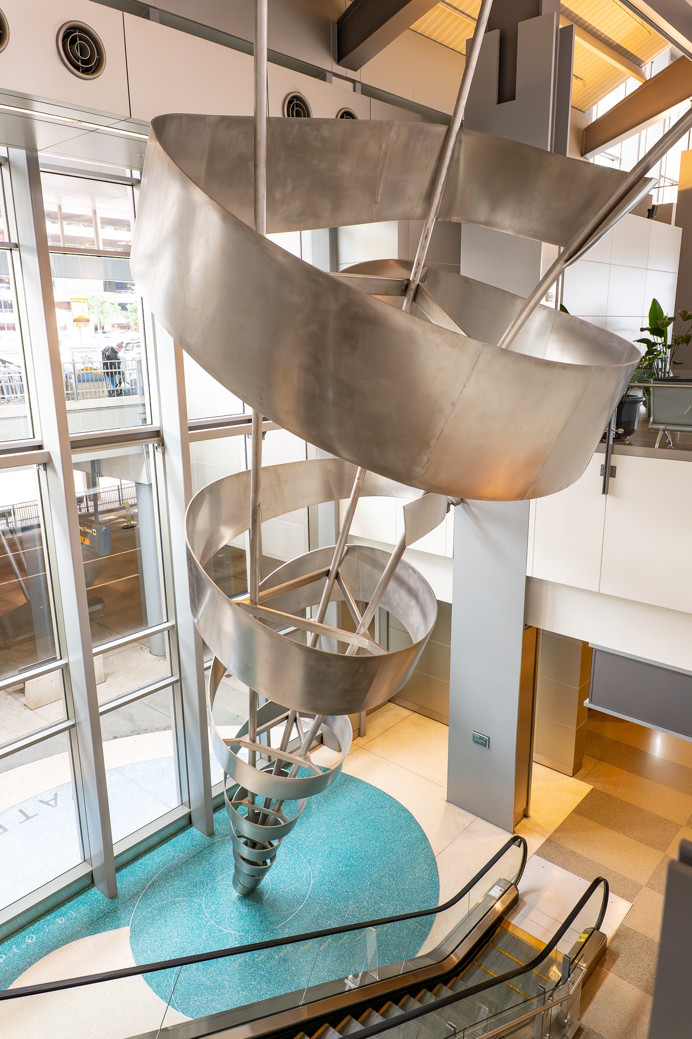 Modern spiral metal sculpture in a bright atrium with glass walls, turquoise floor, and escalators below.