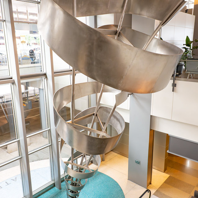 Modern spiral metal sculpture in a bright atrium with glass walls, turquoise floor, and escalators below.