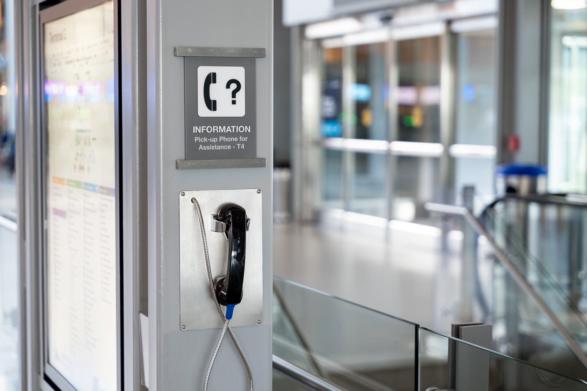 Information assistance phone mounted on gray pillar in modern airport terminal with glass railings and escalators.