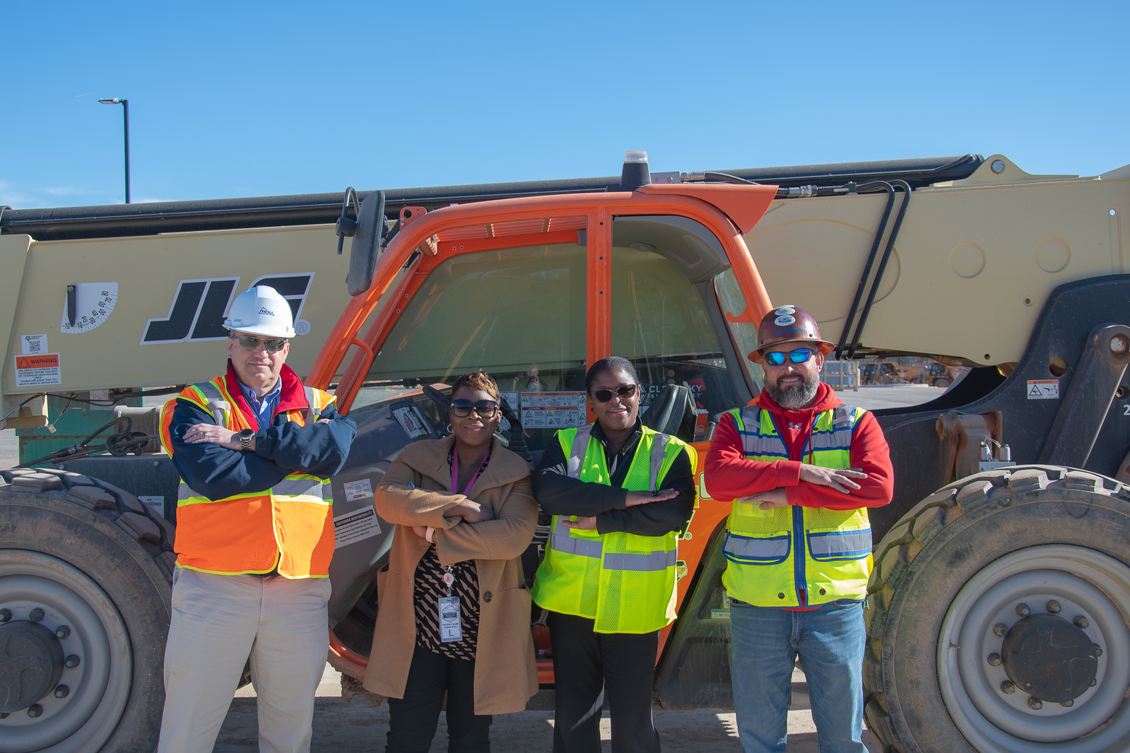 Four Airport Authority employees in safety vests and hard hats stand with arms crossed in front of an orange excavator.
