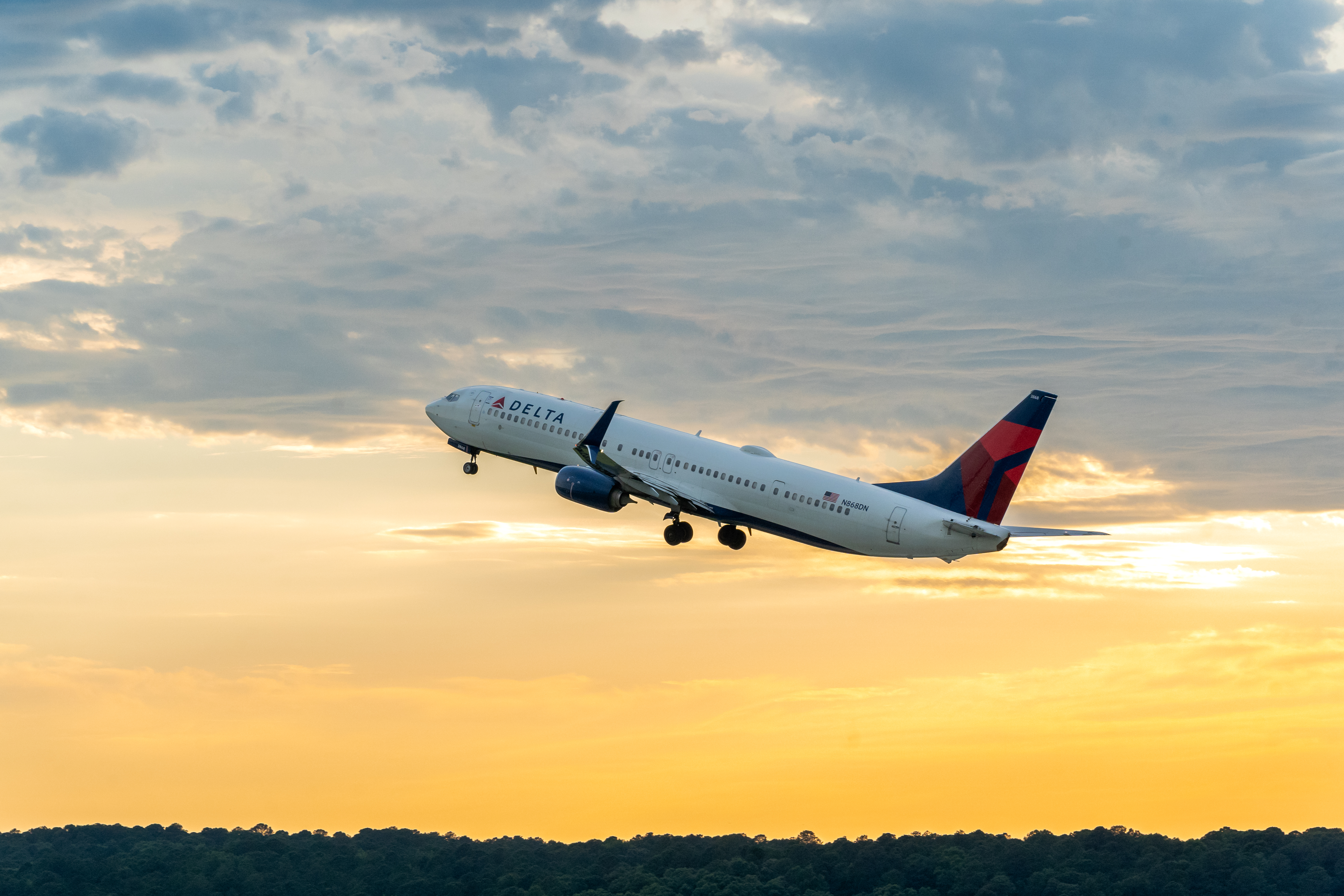 Delta airplane taking off against a dramatic sunset sky with golden and blue hues, silhouetted trees below.