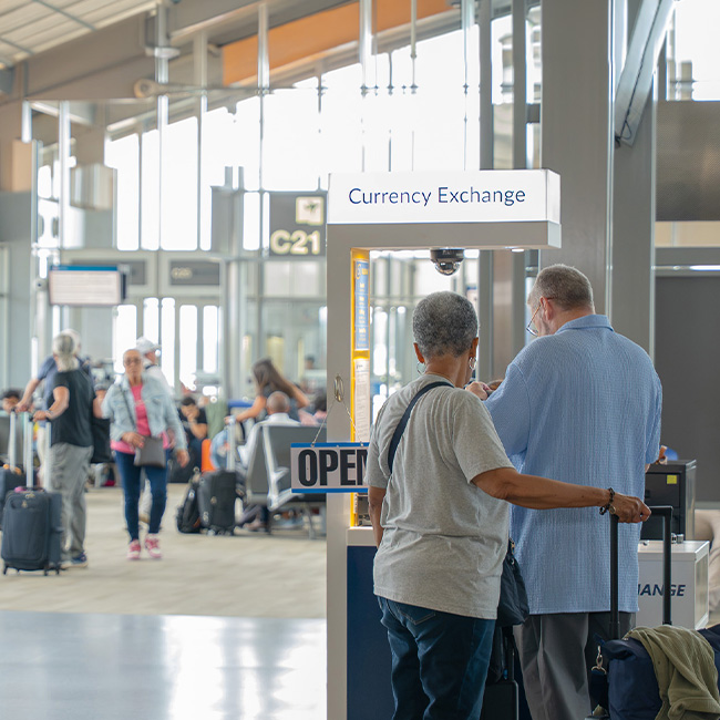 Two travelers with luggage stand at an open currency exchange kiosk in a busy airport terminal with gate signs visible.