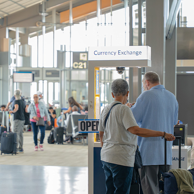 Two travelers with luggage stand at an open currency exchange kiosk in a busy airport terminal with gate signs visible.