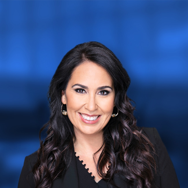 Professional headshot of a smiling woman with long dark wavy hair wearing a black top and gold earrings against a blue background.
