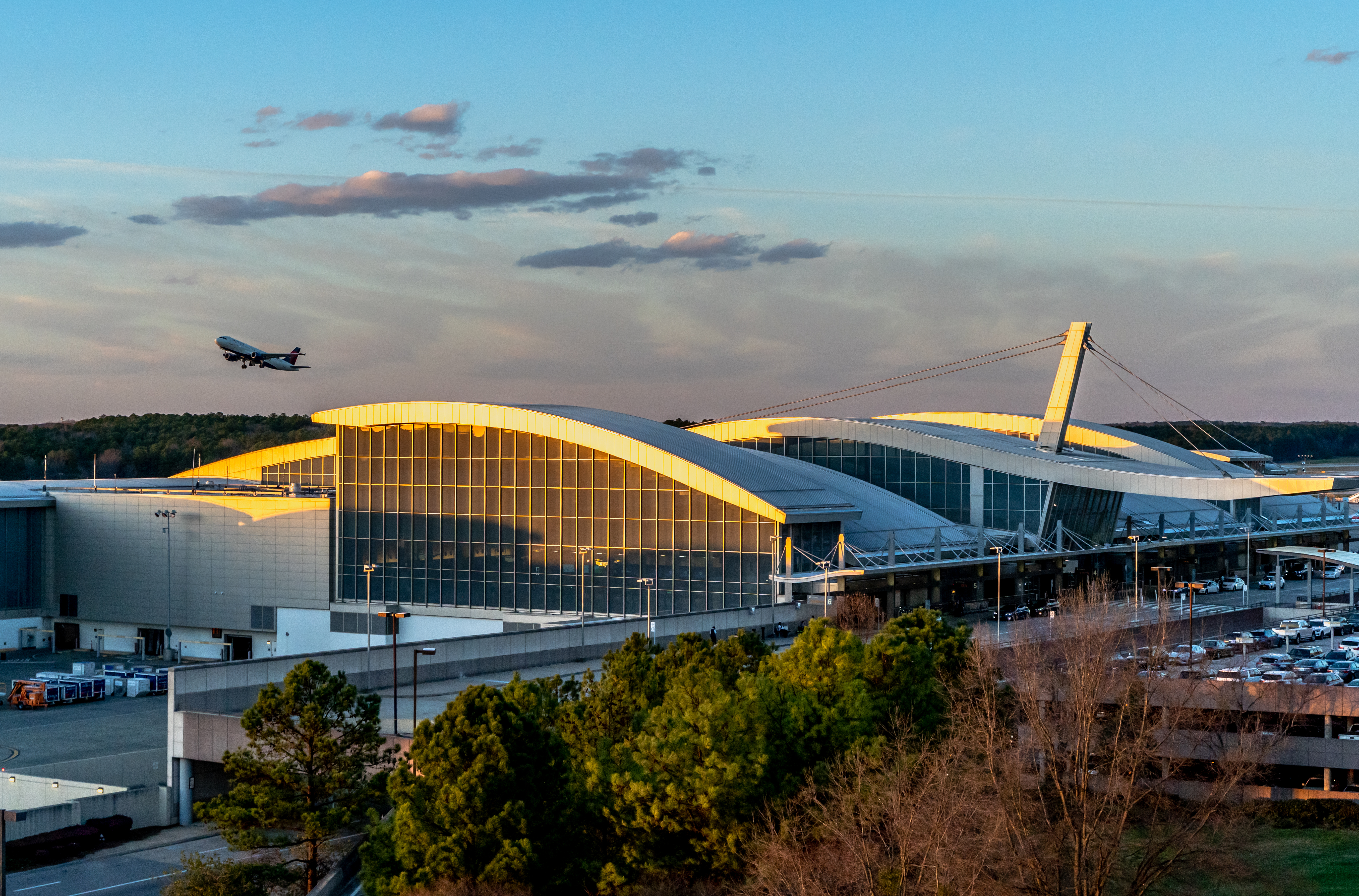 Raleigh-Durham airport terminal two with curved white roof and glass facade at sunset, with airplane taking off in blue sky above.