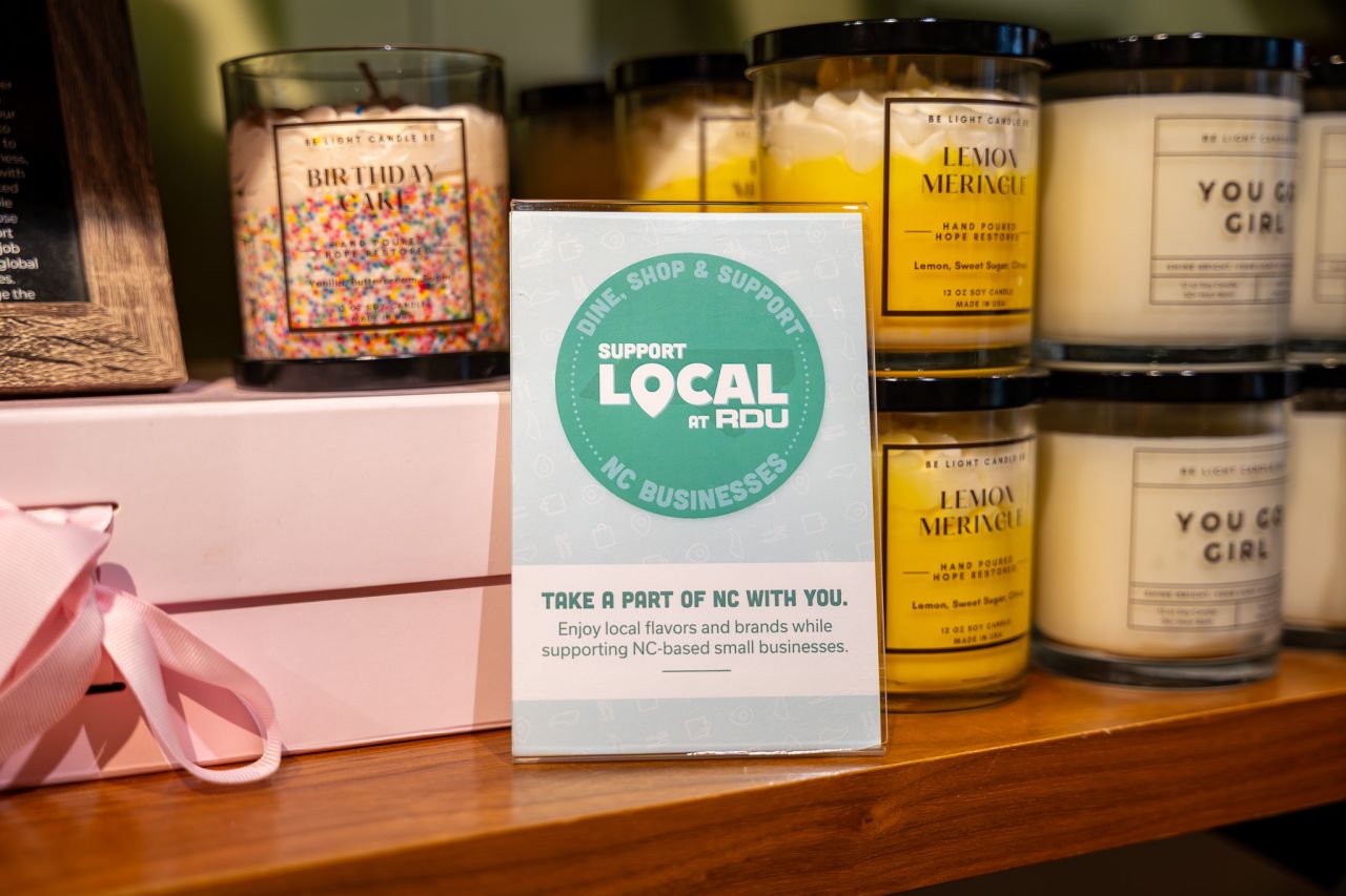 Support Local at RDU sign displayed with scented candles and pink gift box on wooden shelf promoting NC businesses.