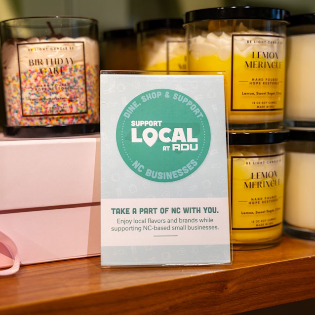 Support Local at RDU sign displayed with scented candles and pink gift box on wooden shelf promoting NC businesses.
