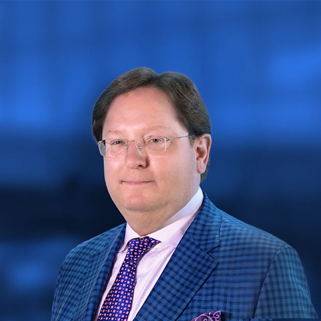 Professional headshot of a man in glasses wearing a blue checkered suit jacket and patterned tie against a blue background.