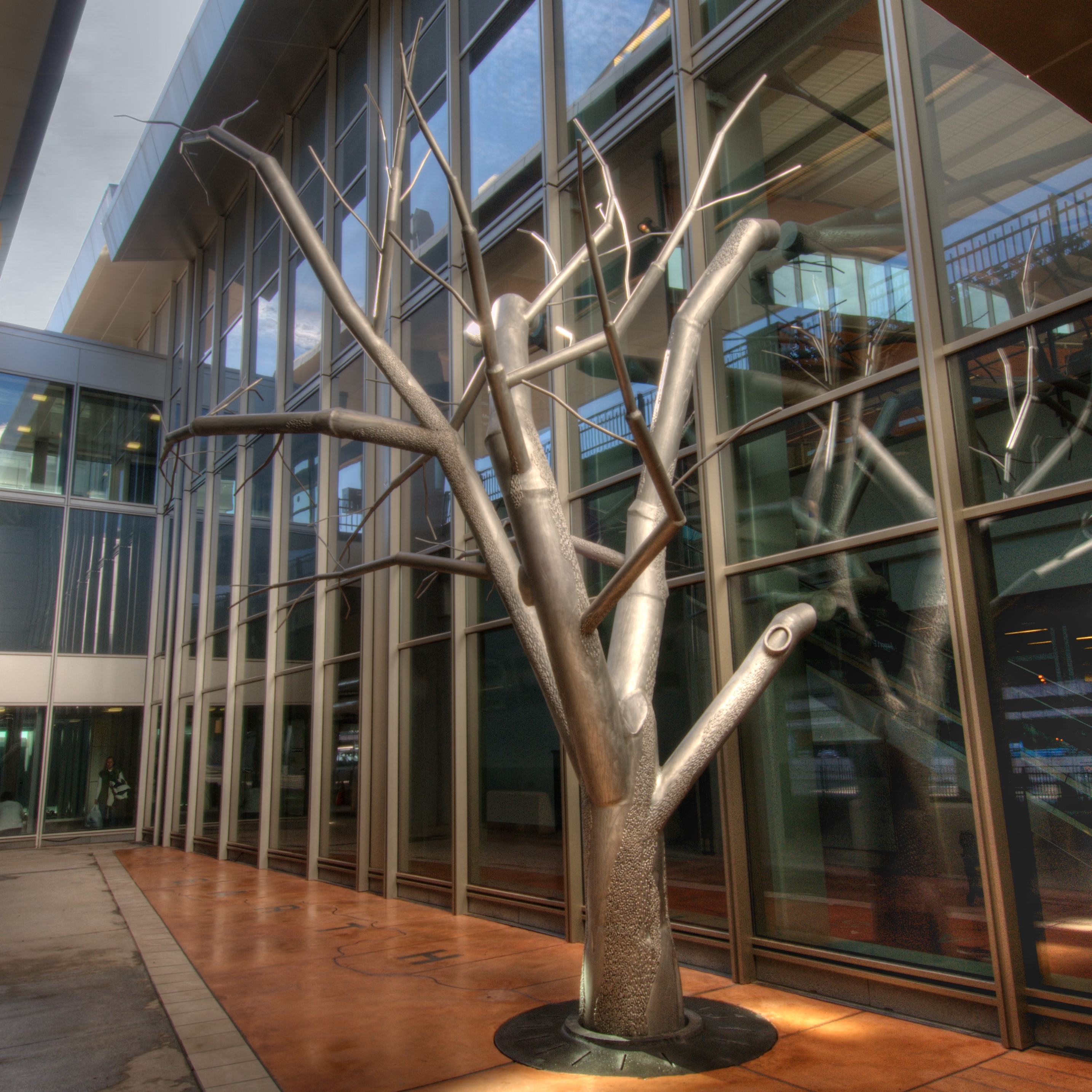 Modern courtyard with metallic tree sculpture on orange-tiled floor, surrounded by glass walls and overhangs.