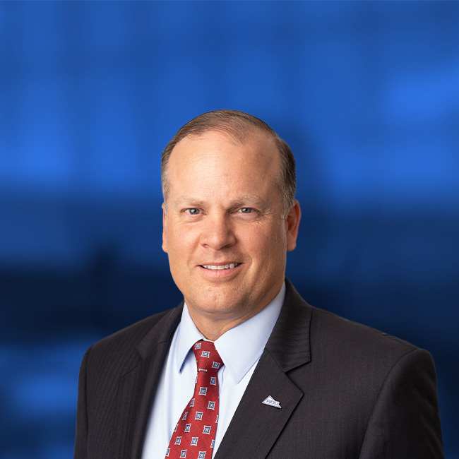 Professional headshot of a smiling man in a dark suit with red patterned tie and white shirt against a blue background.