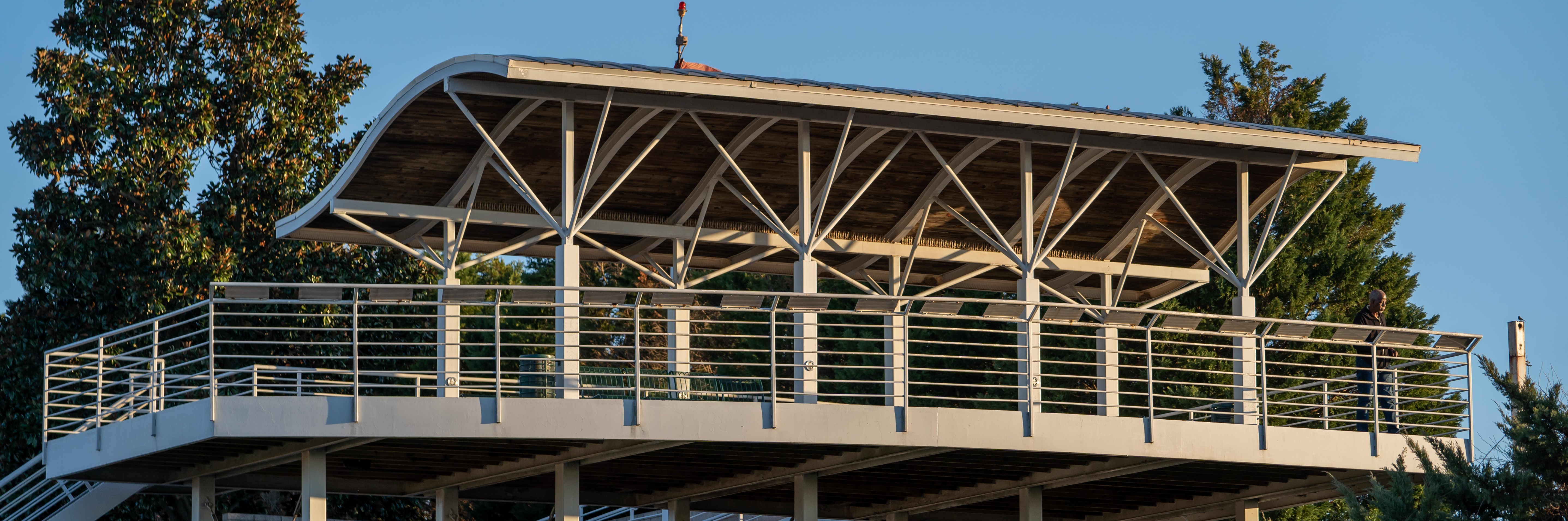 Modern pavilion with curved roof and diagonal support beams, elevated on pillars with metal railing, against blue sky.