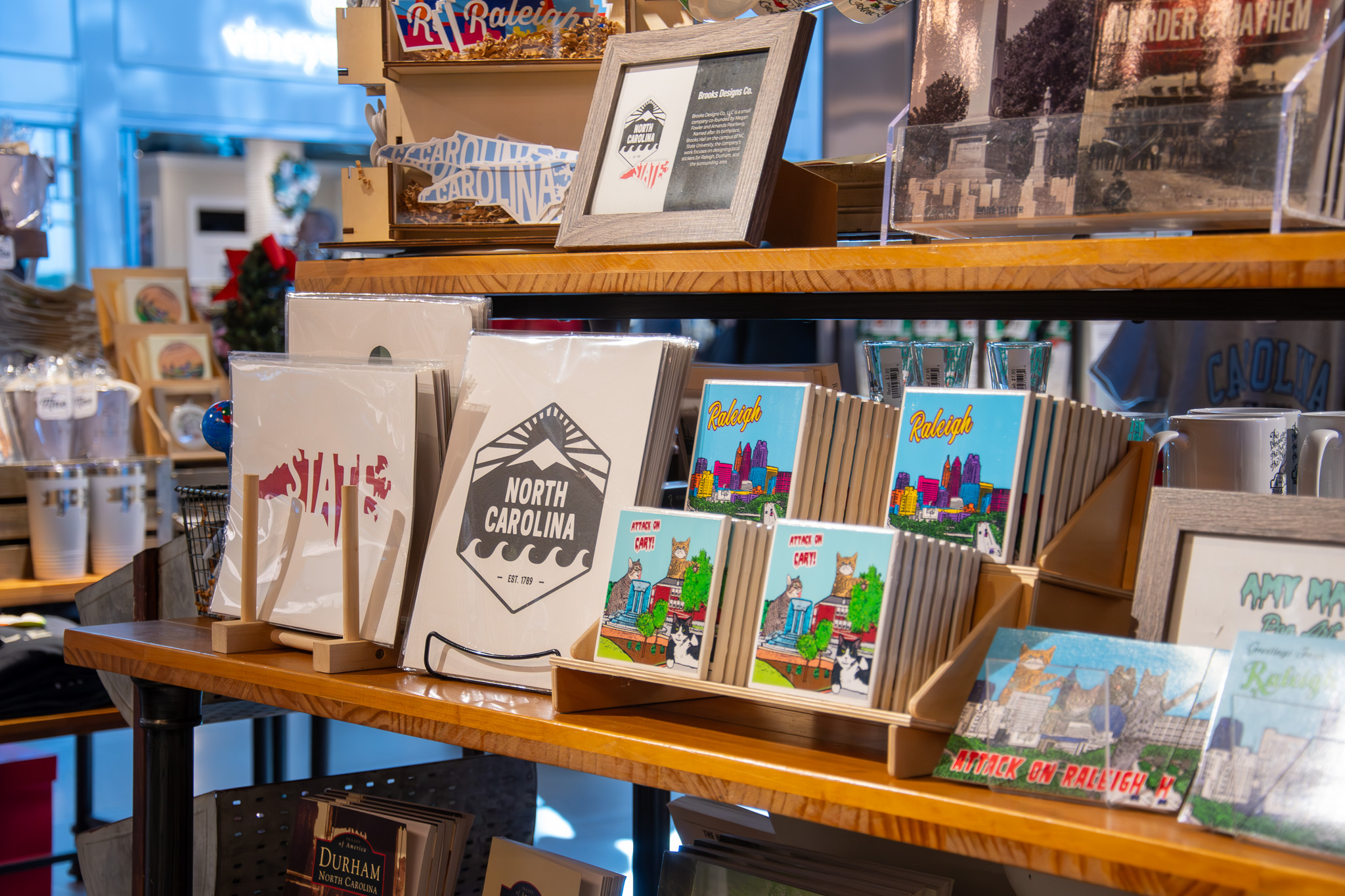 Wooden shelves displaying North Carolina-themed greeting cards, prints, and souvenirs featuring Raleigh skyline and local landmarks.