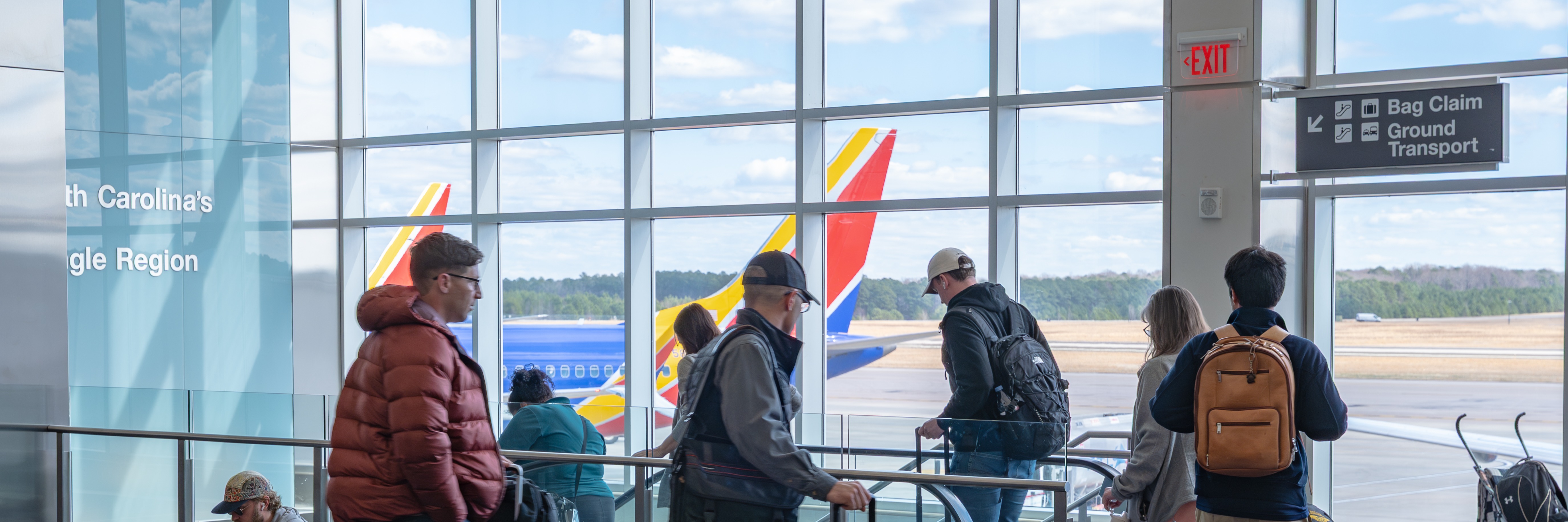 Travelers with luggage walking through modern airport terminal with large windows showing airplane on tarmac outside.