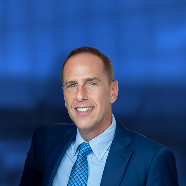 Professional headshot of a smiling man in a navy suit, light blue shirt, and blue checkered tie against a blue background.