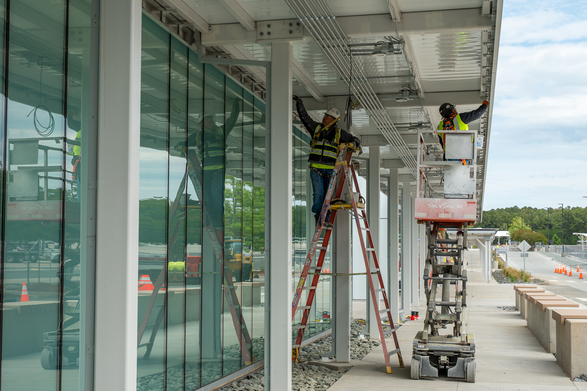 Construction workers in safety vests, one on a ladder and another in a cherry picker lift, work on RDU's new Customer Amenity Building at ParkRDU Economy 3.