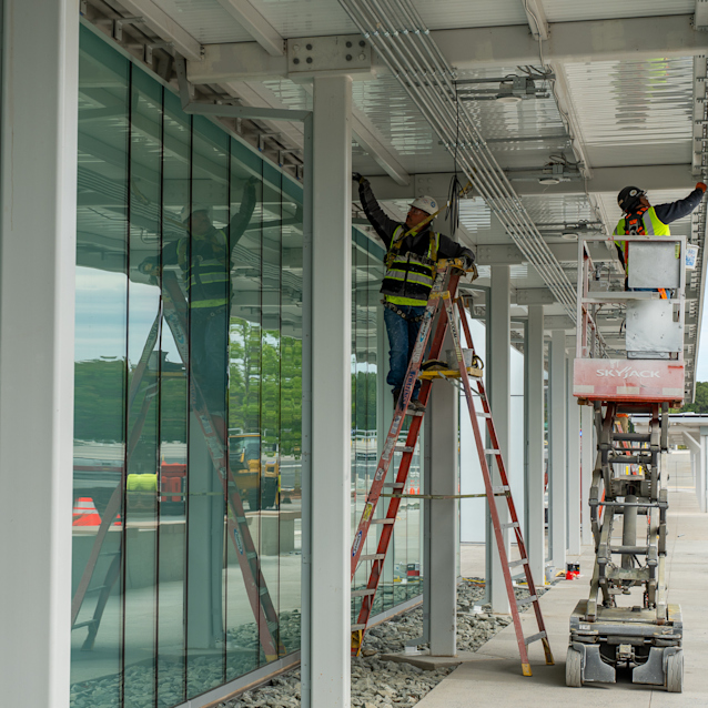 Construction workers in safety vests, one on a ladder and another in a cherry picker lift, work on RDU's new Customer Amenity Building at ParkRDU Economy 3.