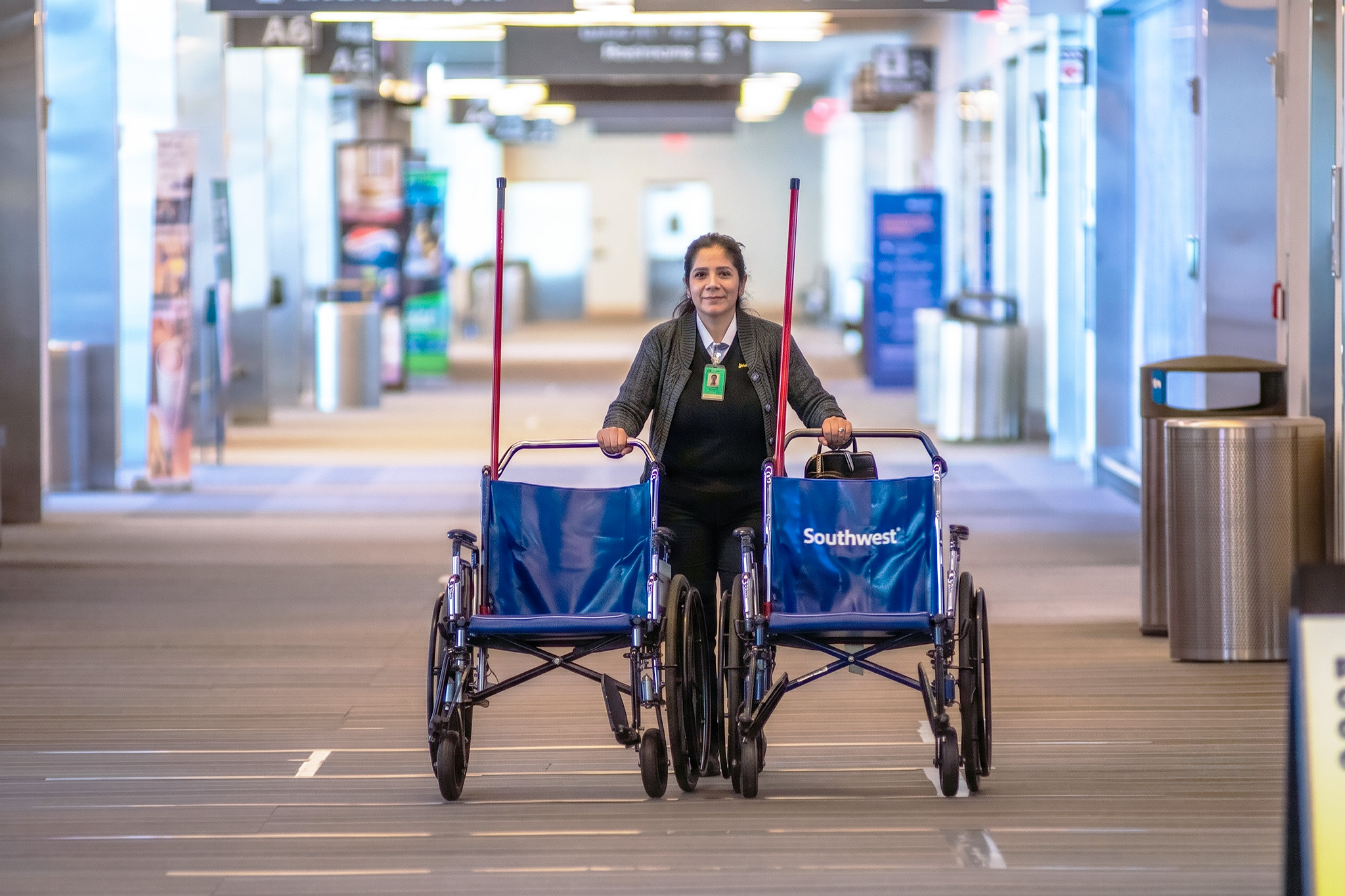 Airport employee pushing two blue Southwest-branded wheelchairs through a bright terminal corridor.