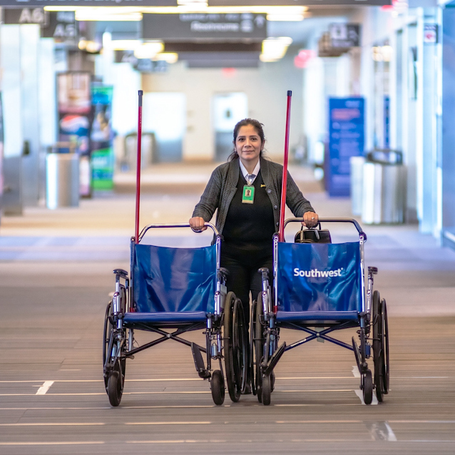 Airport employee pushing two blue Southwest-branded wheelchairs through a bright terminal corridor.