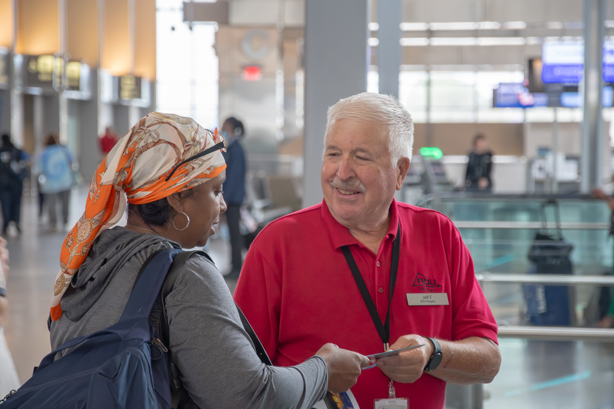 Airport volunteer staff member in red polo shirt assisting a traveler wearing an orange headscarf in a modern terminal.