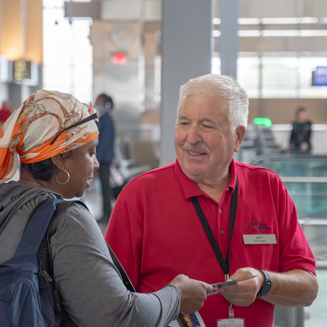 Airport volunteer staff member in red polo shirt assisting a traveler wearing an orange headscarf in a modern terminal.