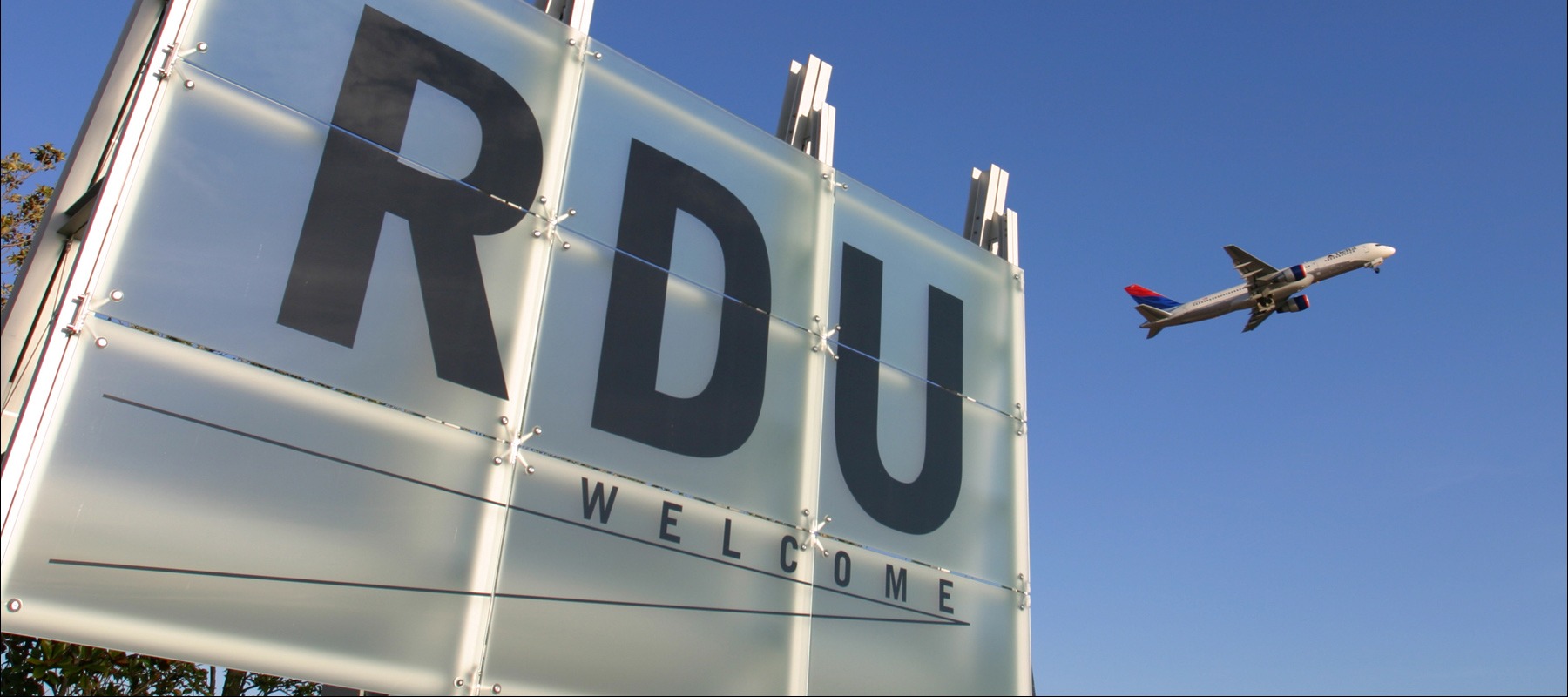White RDU Welcome sign with airplane flying overhead against clear blue sky at Raleigh-Durham International Airport.