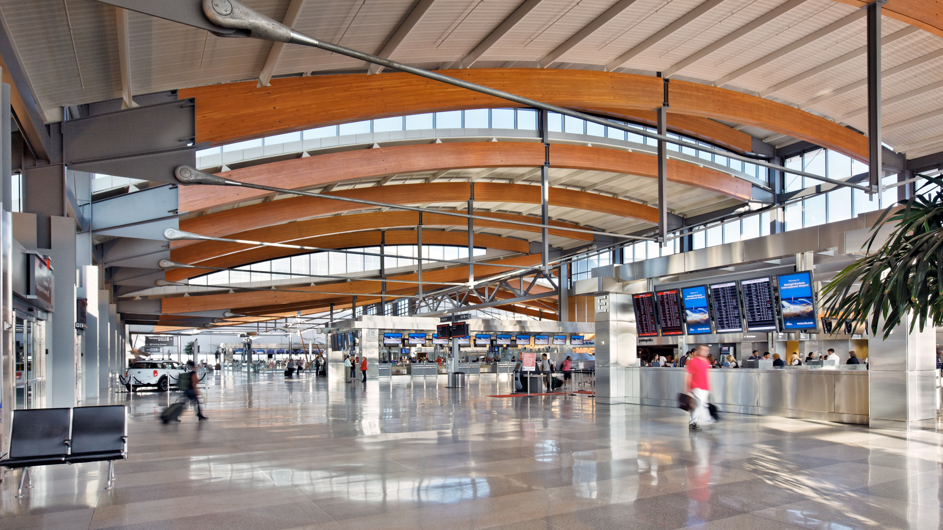 Modern airport terminal with curved wooden beams, high ceilings, large windows, and travelers walking through the spacious hall.