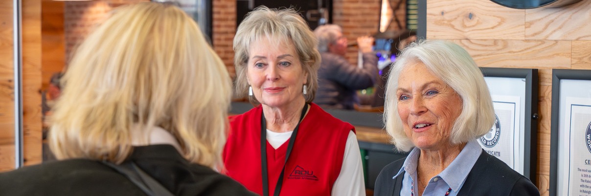 Three women conversing in a modern office with brick walls and wooden panels, one wearing a red vest, another in blue.