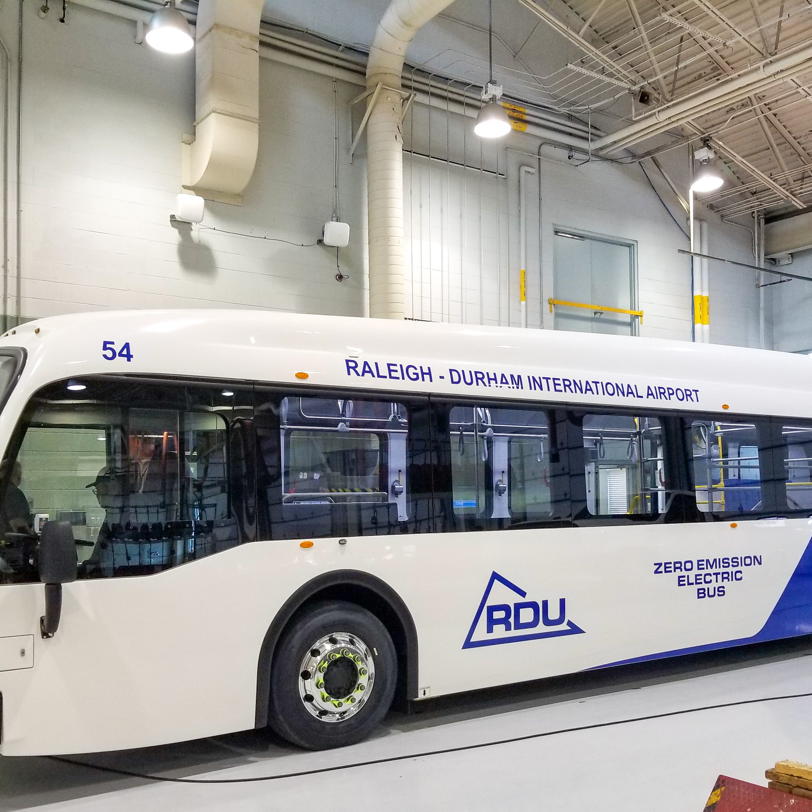 White electric bus labeled Raleigh-Durham International Airport and RDU parked inside an industrial facility.