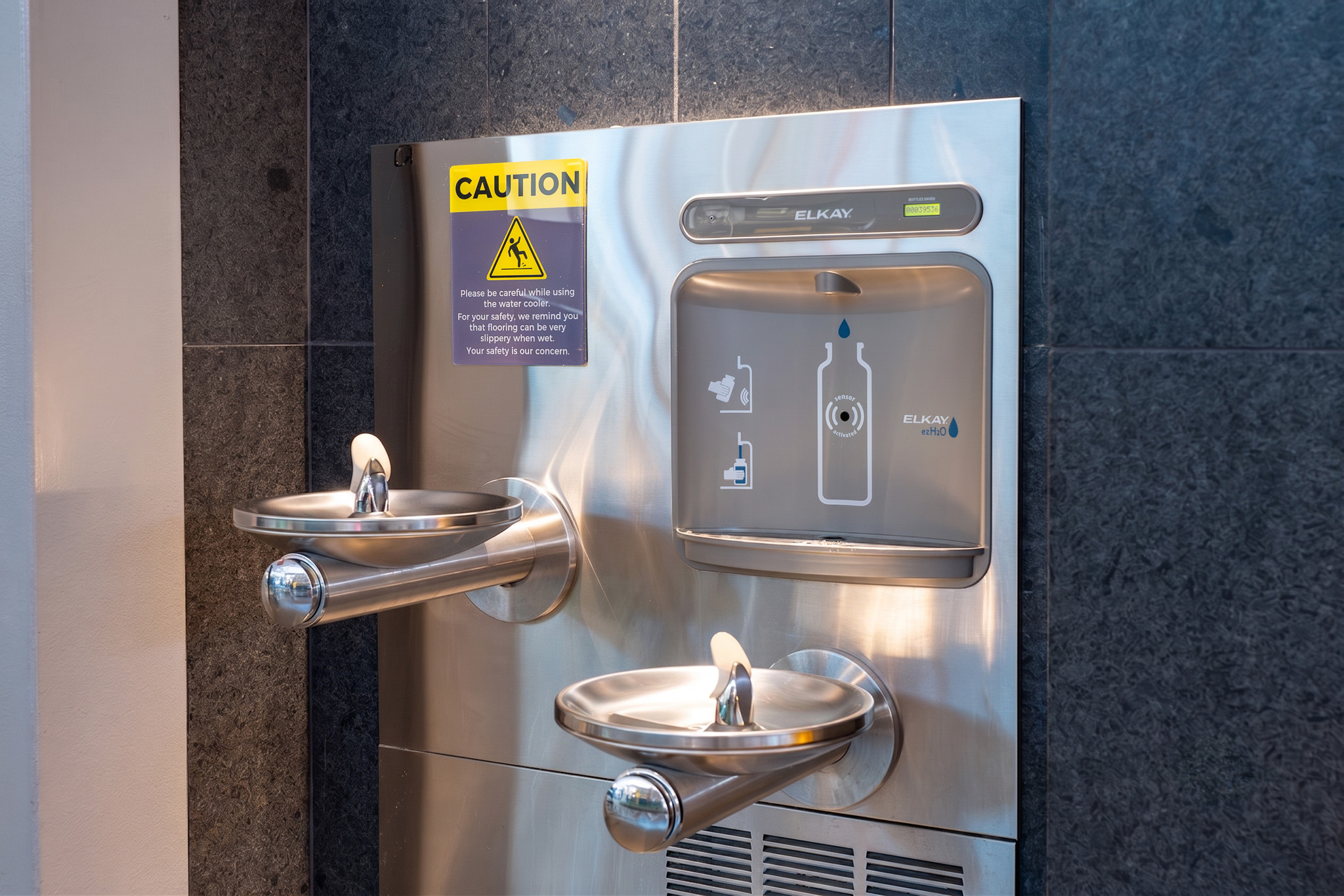 Stainless steel water fountain and bottle filling station mounted on dark tiled wall with yellow caution sign above.