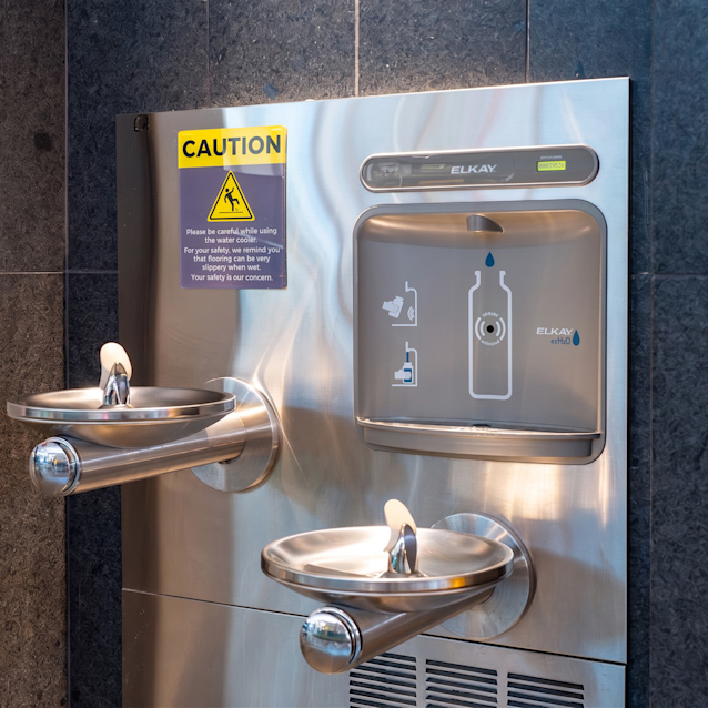 Stainless steel water fountain and bottle filling station mounted on dark tiled wall with yellow caution sign above.