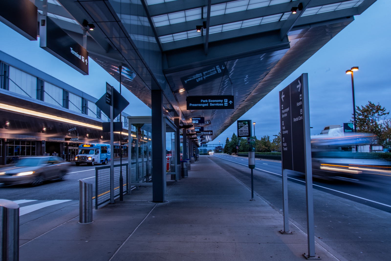 Terminal 1 exterior at dusk with illuminated canopy, directional signs, and vehicles in motion on roadway.
