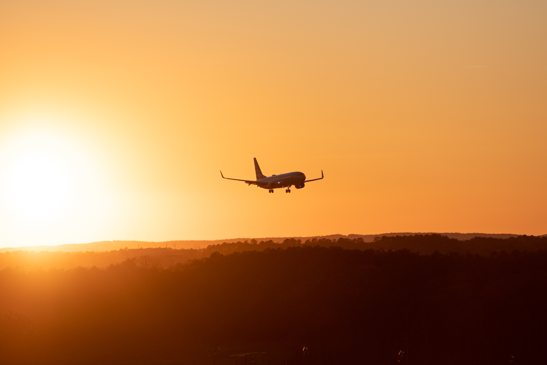 Commercial airplane silhouetted against an orange sunset sky, approaching for landing above dark hills.