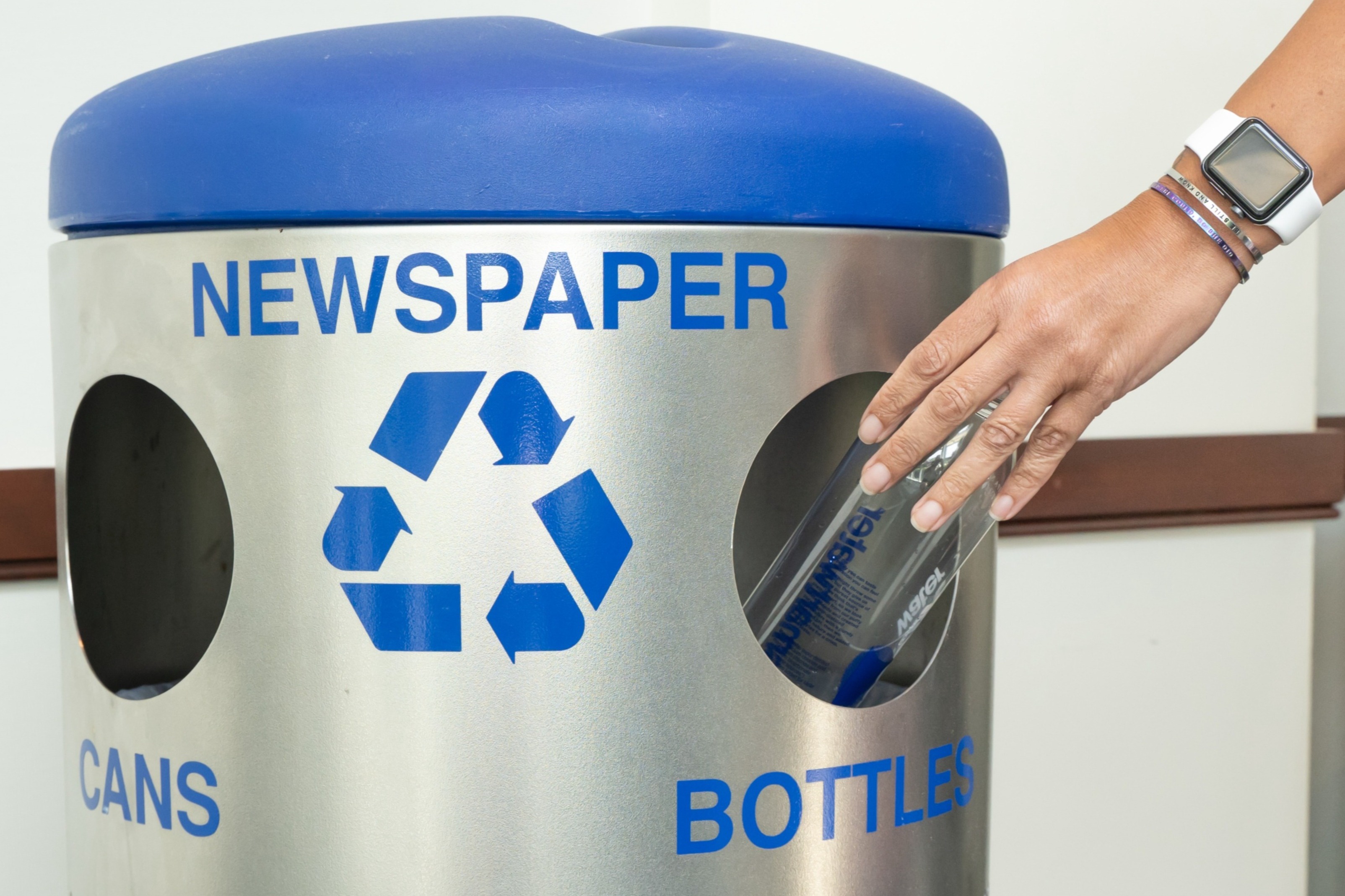 Person placing a plastic bottle into a silver recycling bin with blue lid labeled for newspaper, cans, and bottles.