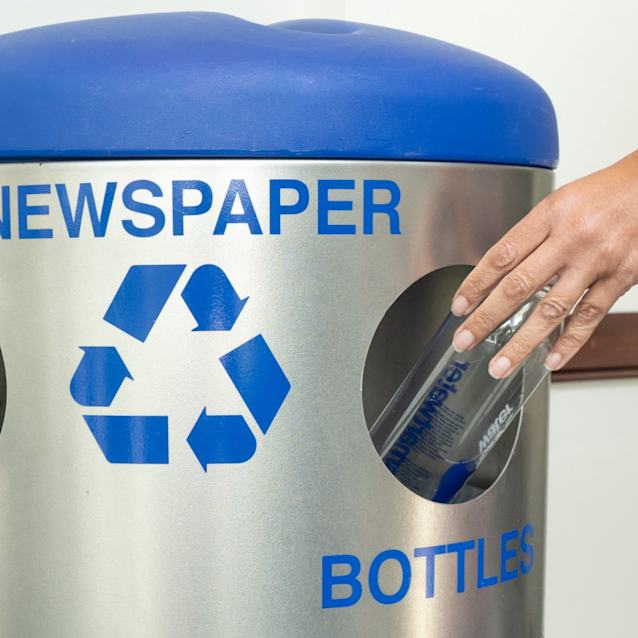 Person placing a plastic bottle into a silver recycling bin with blue lid labeled for newspaper, cans, and bottles.