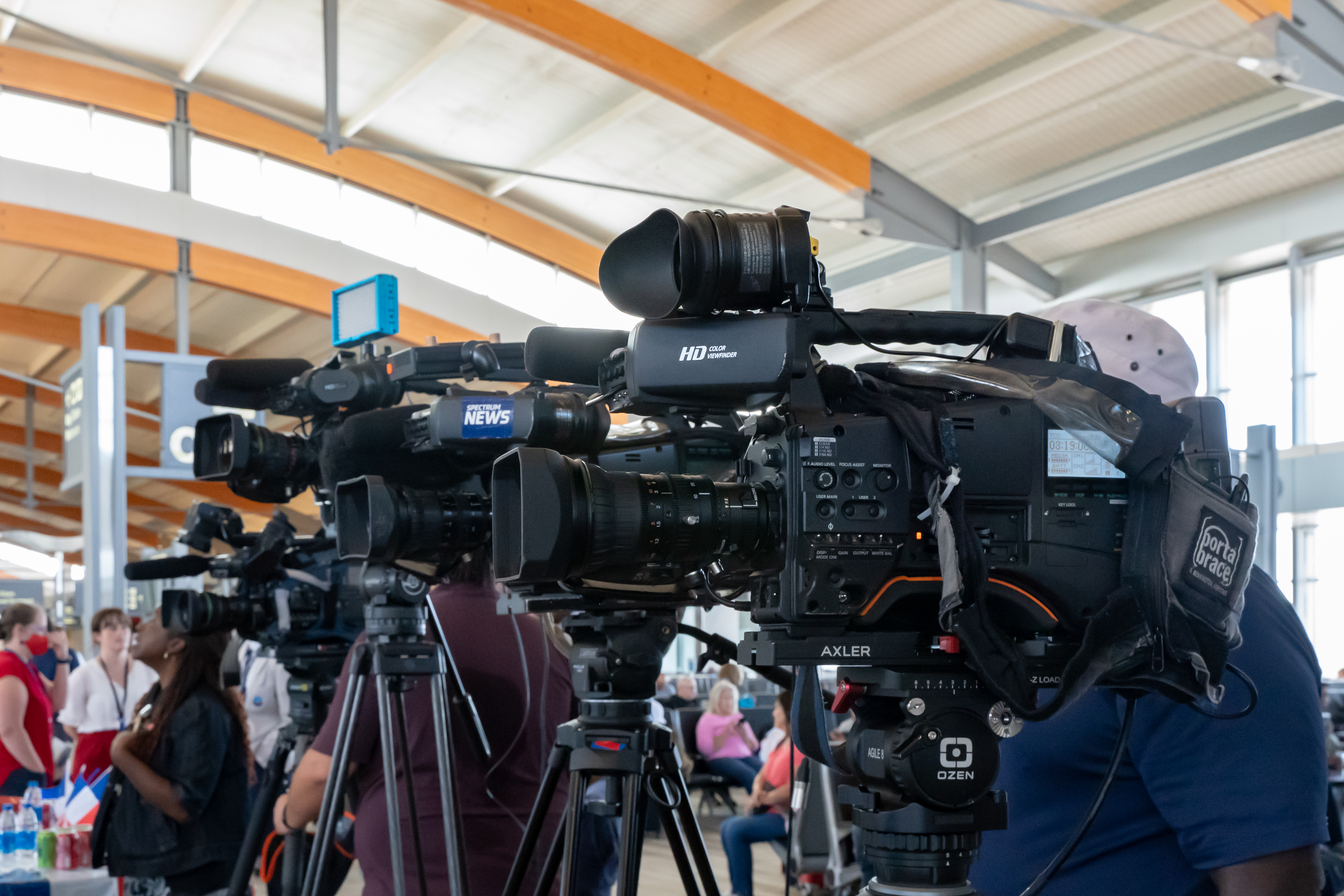 News cameras set up on tripods at an indoor press event, with people sitting and standing in the background.
