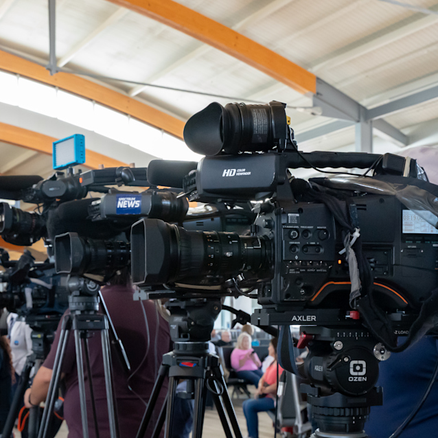 News cameras set up on tripods at an indoor press event, with people sitting and standing in the background.