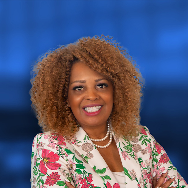 Woman with curly brown hair smiling, wearing a white floral blazer with pink and green flowers and a pearl necklace on blue background.