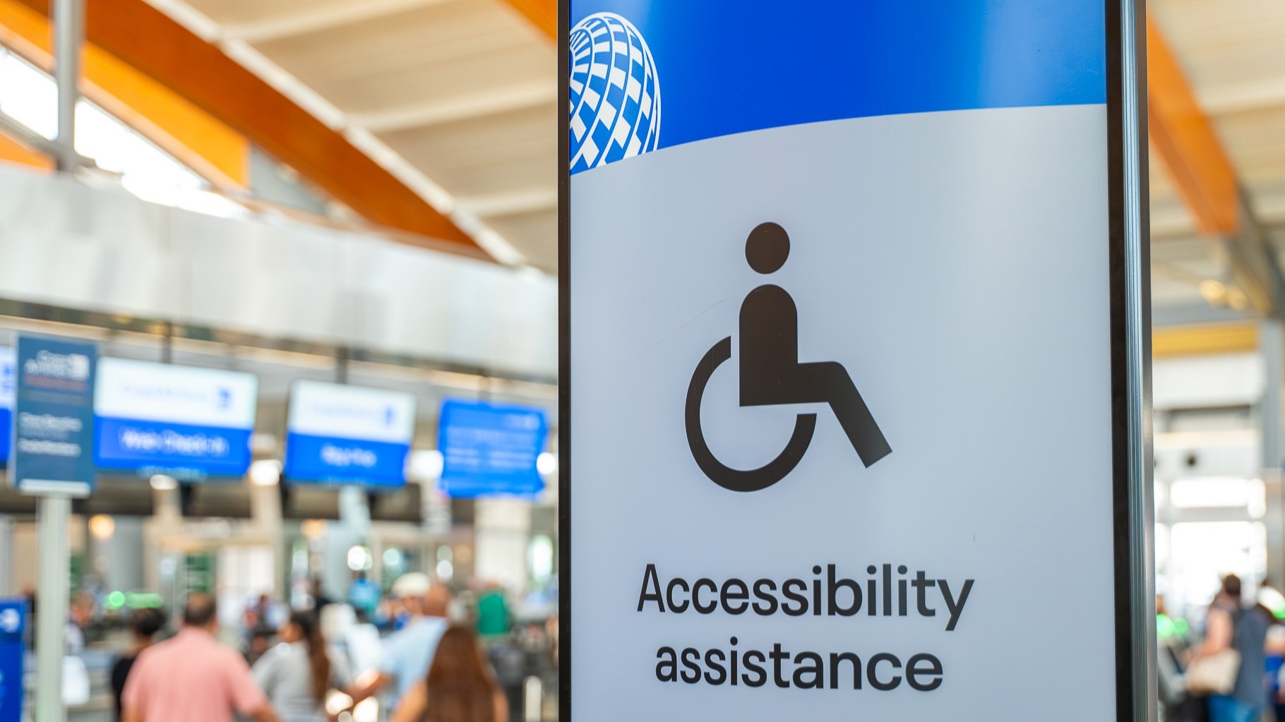 Airport accessibility assistance sign with wheelchair symbol and airline logo in terminal with travelers in background.
