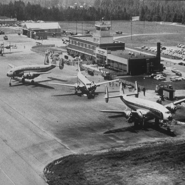Black and white aerial view of vintage propeller aircraft parked on tarmac in front of a mid-century RDU airport terminal building.