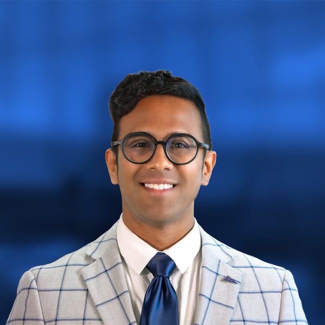 Professional headshot of a smiling man wearing round glasses, a blue tie, and a light gray checkered suit against a blue background.