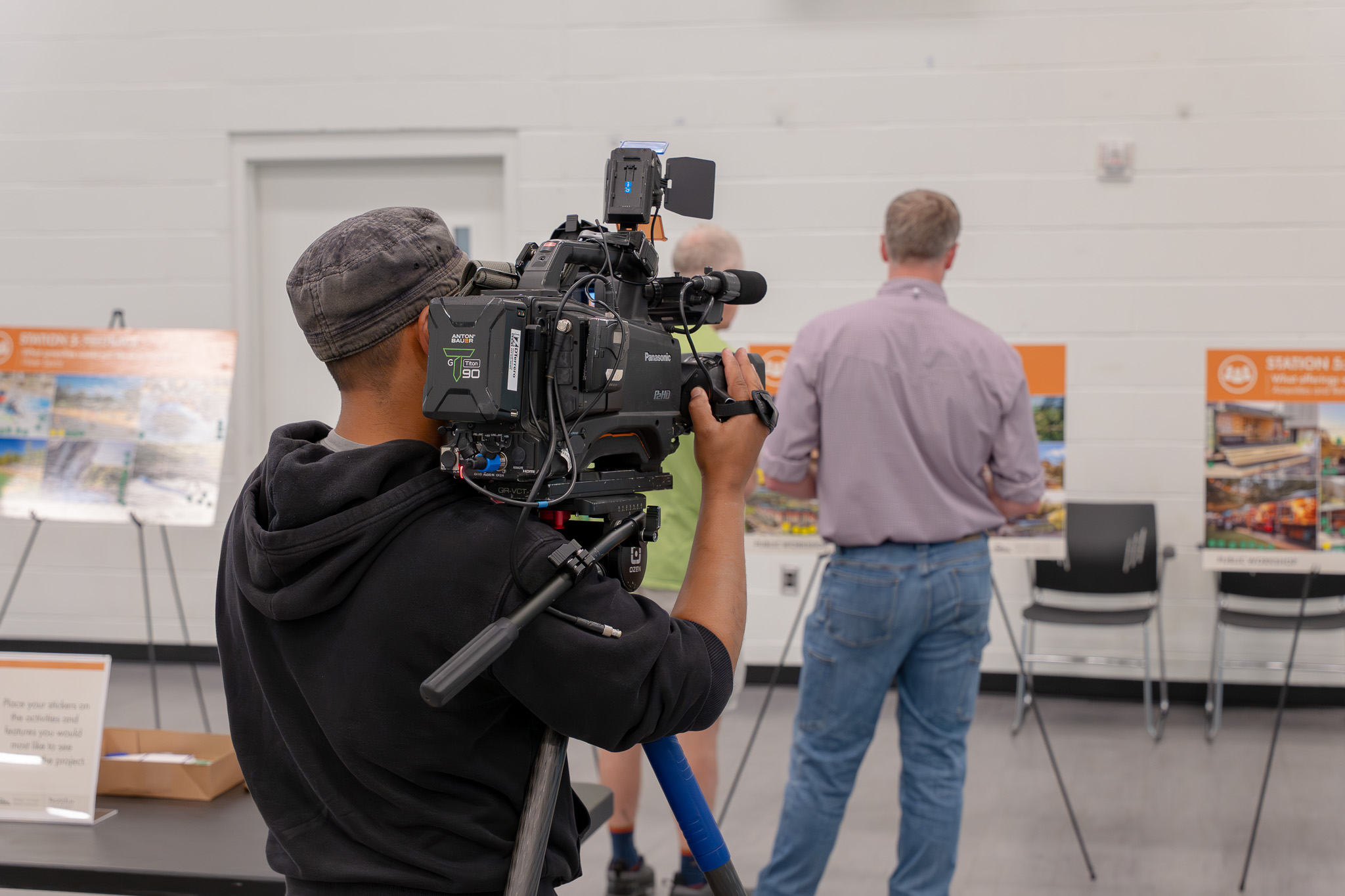 A person is operating a Panasonic video camera on a tripod in a room with white walls. In the background, two people look at informational displays on easels. Chairs are arranged nearby.
