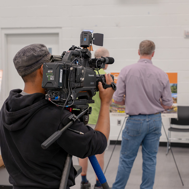 A person is operating a Panasonic video camera on a tripod in a room with white walls. In the background, two people look at informational displays on easels. Chairs are arranged nearby.